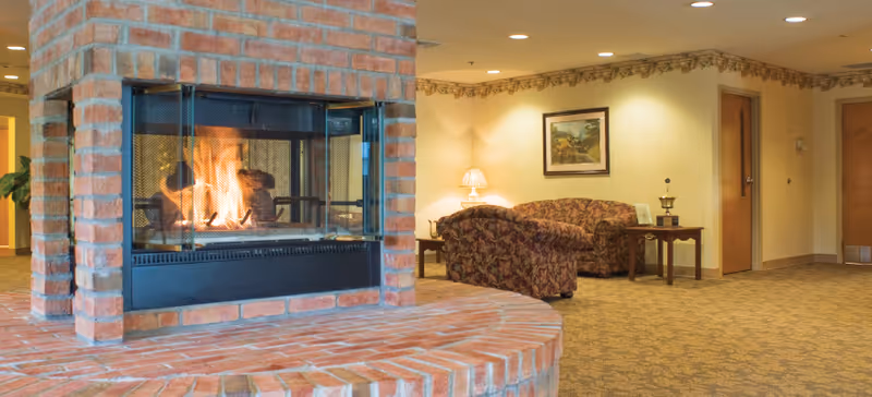 A cozy senior living lounge with a central brick fireplace, patterned sofas, side tables, and warm lighting.
