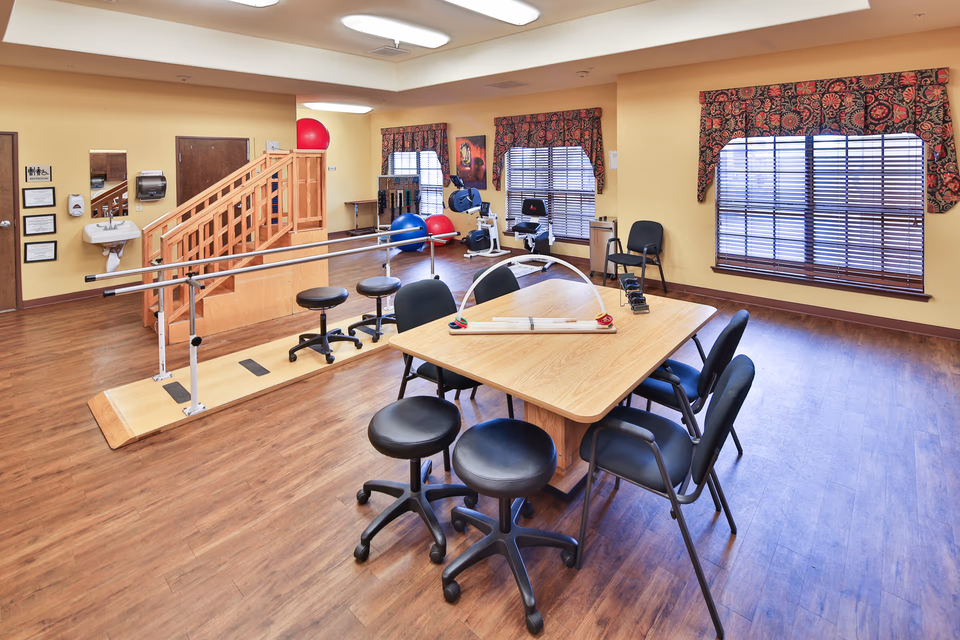 A spacious room with wooden flooring and yellow walls, featuring a wooden table surrounded by black chairs and stools. The room contains physical therapy equipment including parallel bars, exercise balls, and stationary exercise machines. There are three windows with patterned valances and blinds, and a small wooden staircase in the corner.