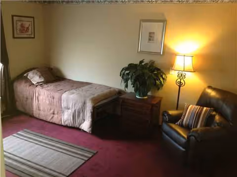 A cozy bedroom in a senior living facility with bed, nightstand, plant, and chair.