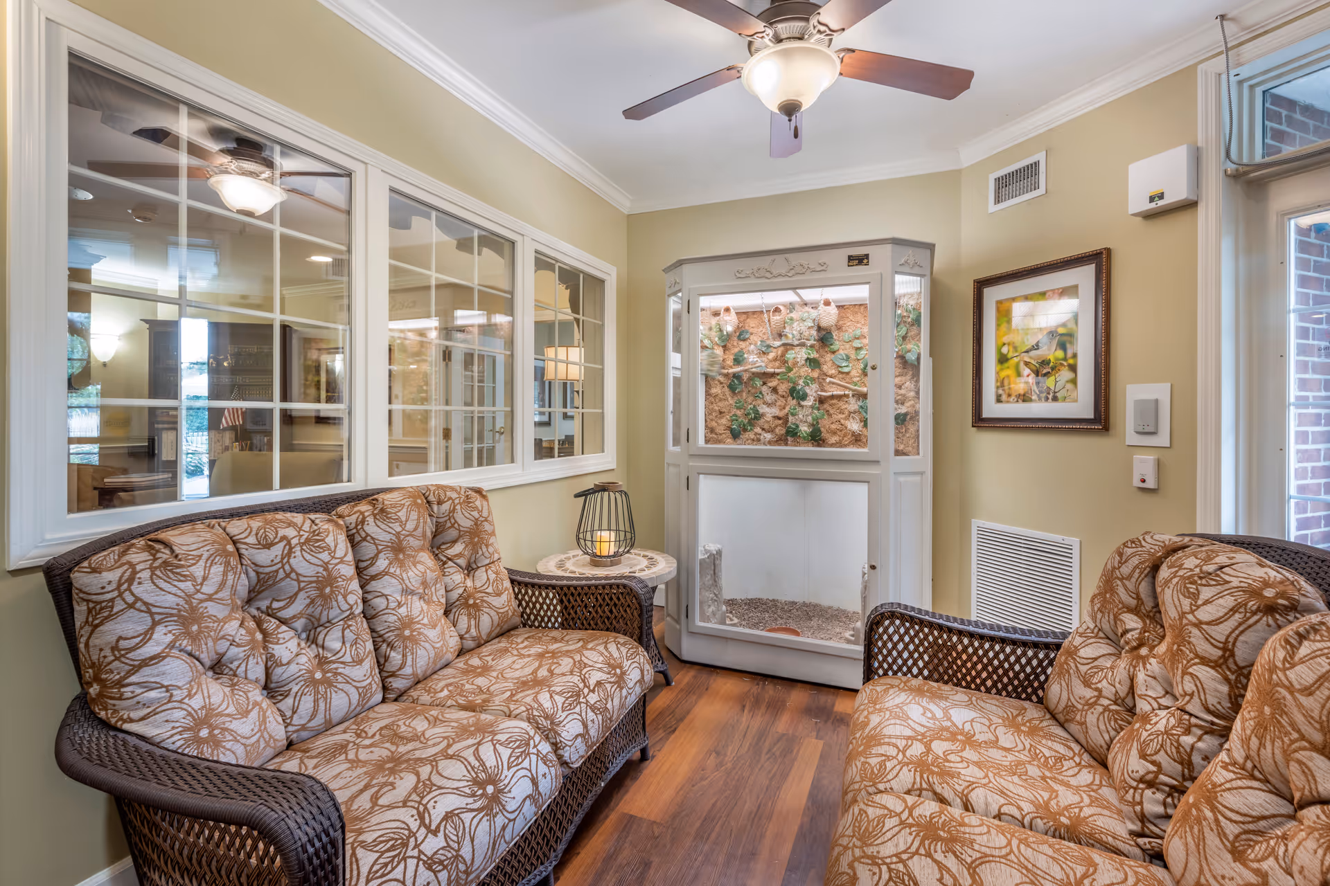 A cozy sitting area in a senior living facility with two cushioned wicker sofas facing each other, a small round table with a decorative candle holder, a ceiling fan with light, and a large glass-enclosed terrarium or bird habitat against the wall. The walls are painted light yellow, and there is a framed picture of a bird on the wall next to a window and door with brick visible outside.