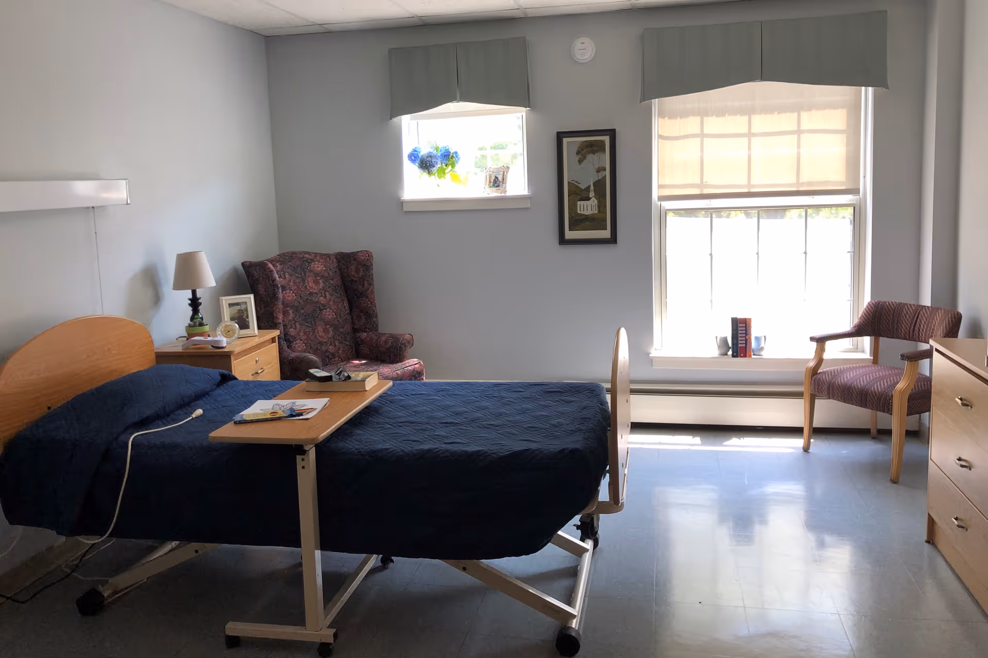 A senior living facility bedroom with a hospital-style bed covered in a dark blue quilt, a wooden bedside table with a lamp and framed photo, a floral upholstered armchair, two windows with light green valances, a framed picture on the wall, a wooden dresser, and a wooden chair near the window.