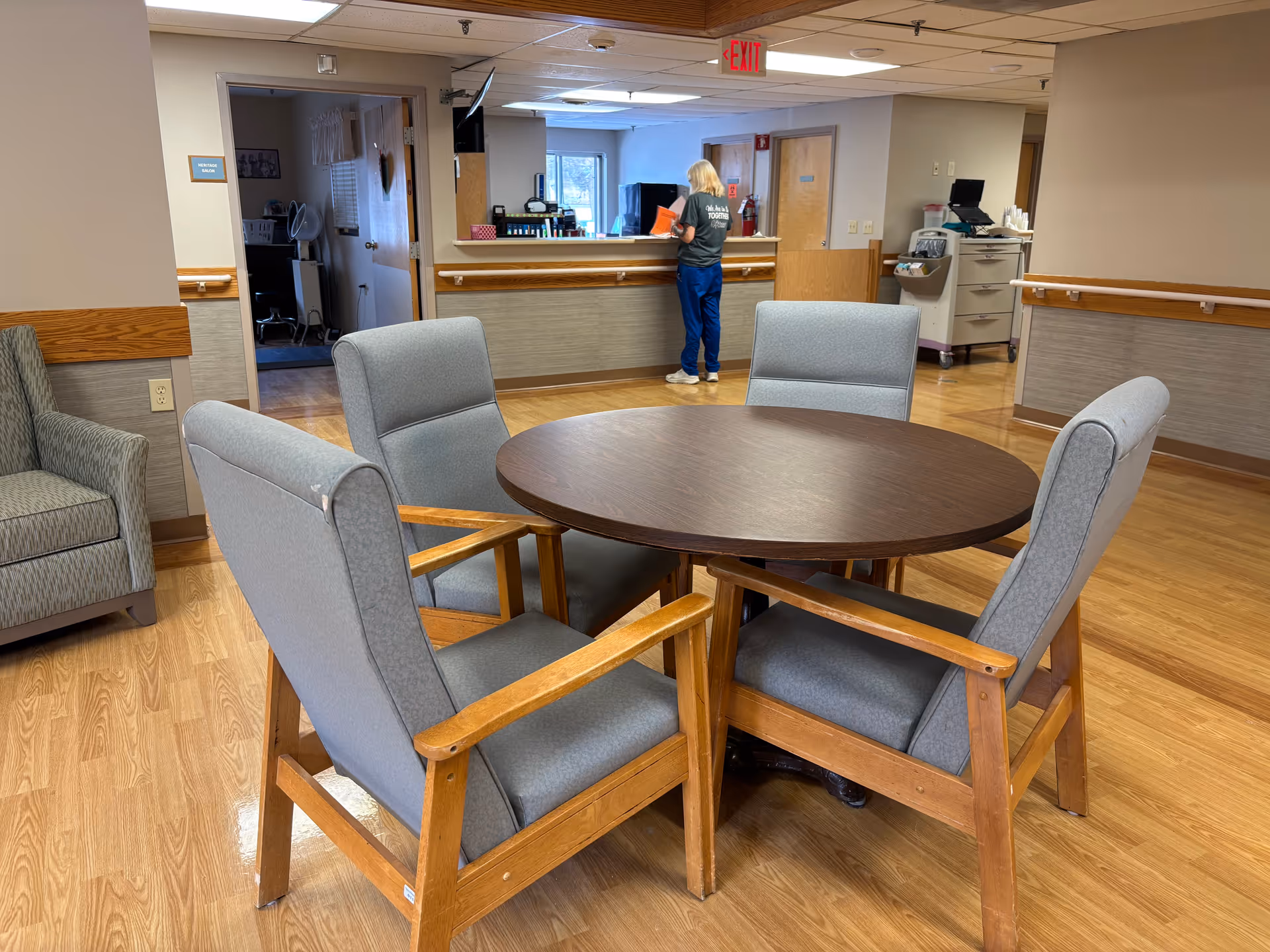 A seating area in a senior living facility with a round wooden table surrounded by four cushioned chairs with wooden arms. In the background, a staff member is standing at a reception or nurse's station counter. The room has wood flooring, beige walls with wooden trim, and an exit sign above a door.