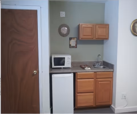 Small kitchenette with wooden cabinets, countertop with a microwave and sink, a mini refrigerator, and a closed wooden door to the left.