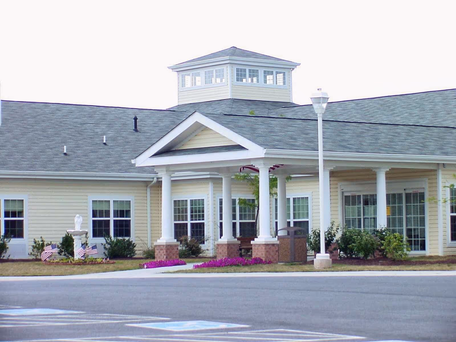 Front entrance of a single-story senior living building with white columns, windows, and landscaped flower beds.