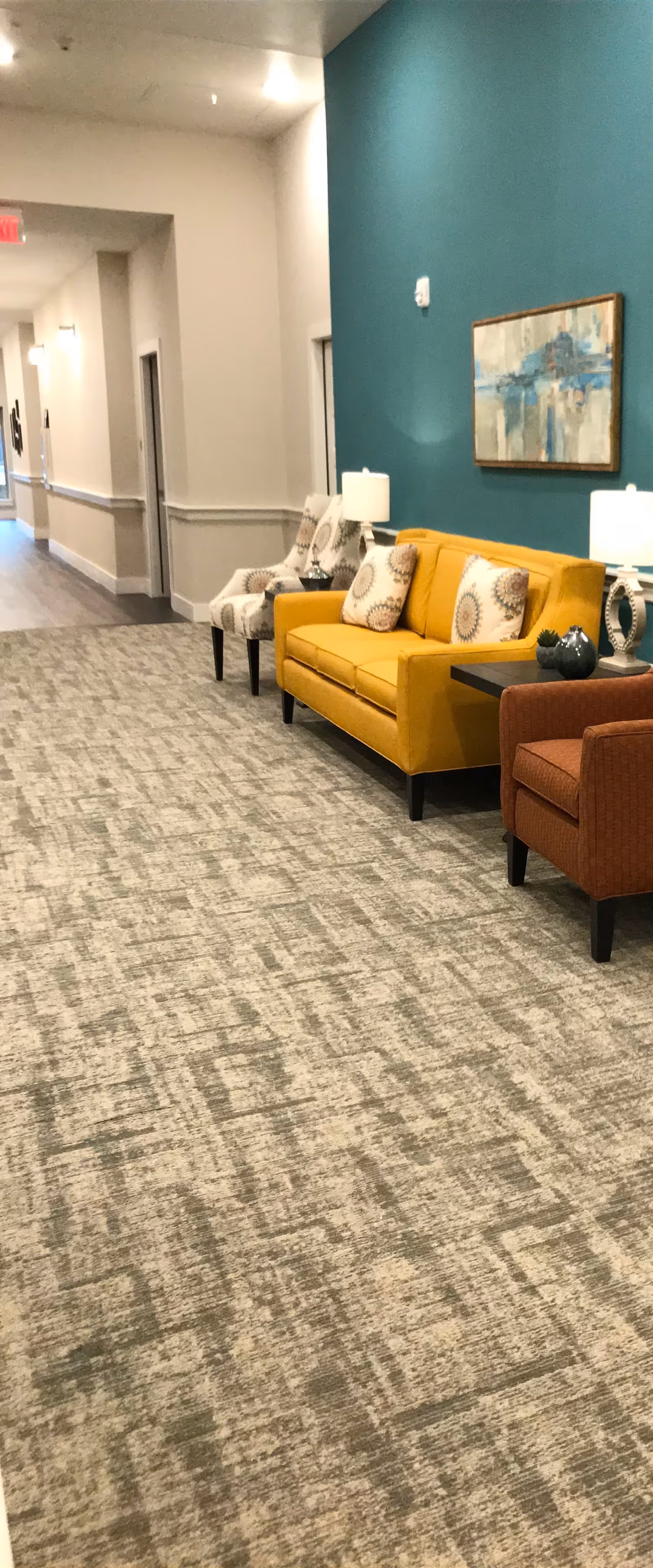 A hallway in an assisted living facility with a patterned carpet floor, a teal accent wall, and seating area including a yellow sofa with decorative pillows, two armchairs, and side tables with lamps. A framed abstract painting hangs on the teal wall.