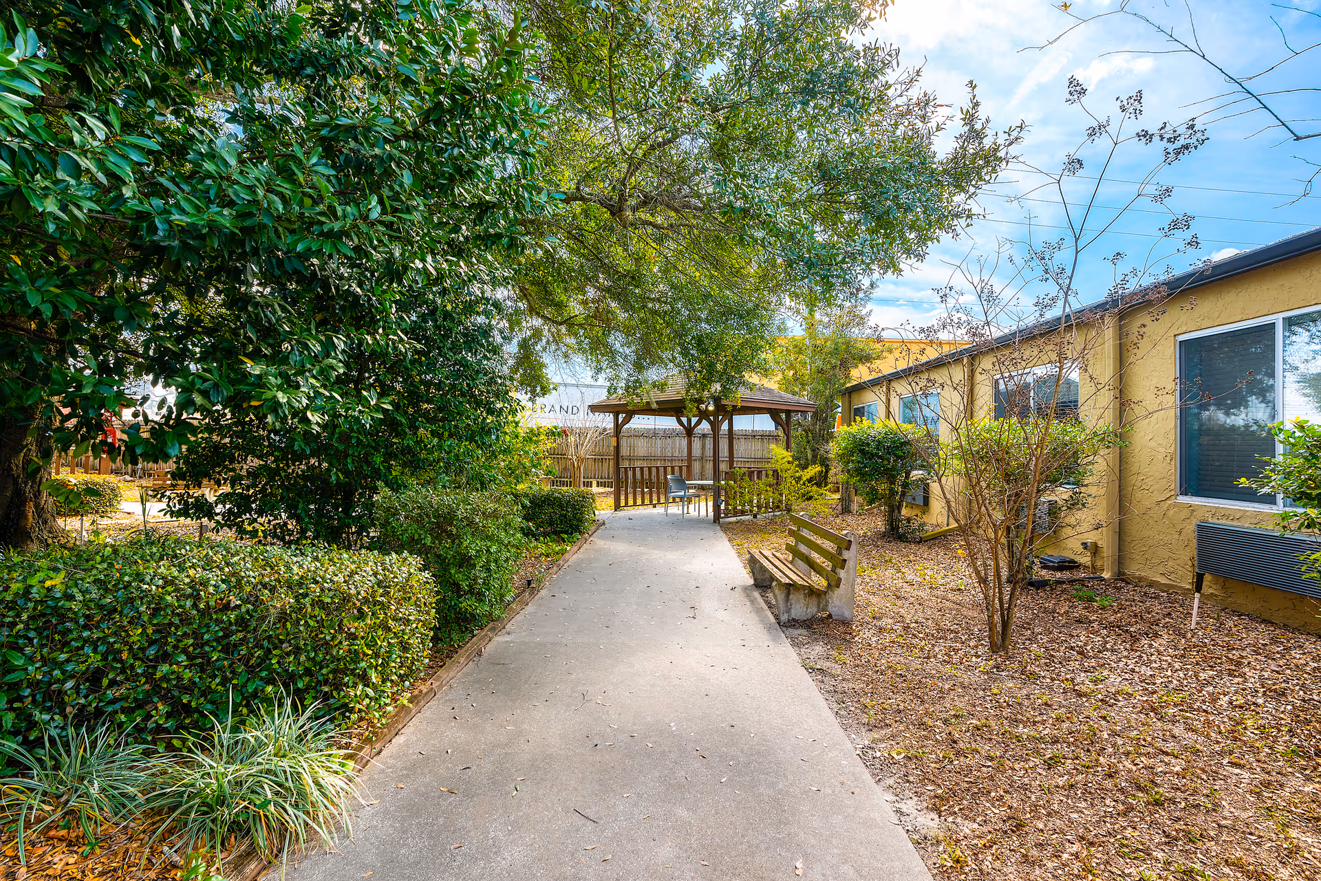 Paved walkway through a landscaped courtyard with benches and a gazebo next to a single-story building.