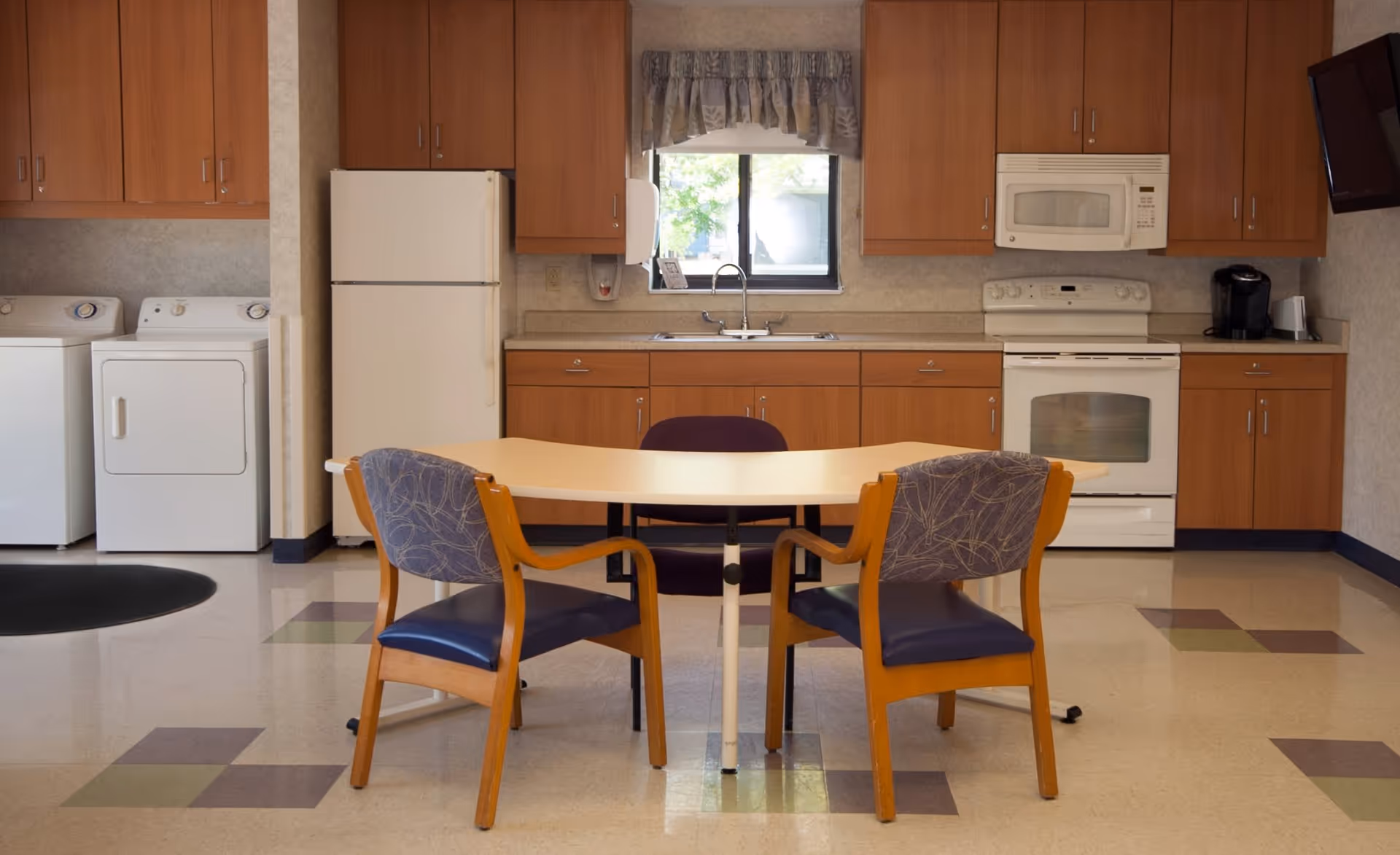 A kitchen area with wooden cabinets, a white refrigerator, stove, microwave, and a coffee maker. In front of the kitchen is a table with three chairs, two with wooden frames and blue patterned upholstery, and one purple chair. To the left, there is a washer and dryer set. A window with a floral valance is above the sink.