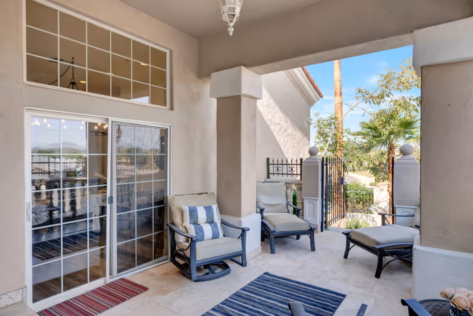 Covered outdoor patio area with cushioned chairs and ottomans, a striped rug, and a view of a gated garden with palm trees and other greenery under a clear blue sky.