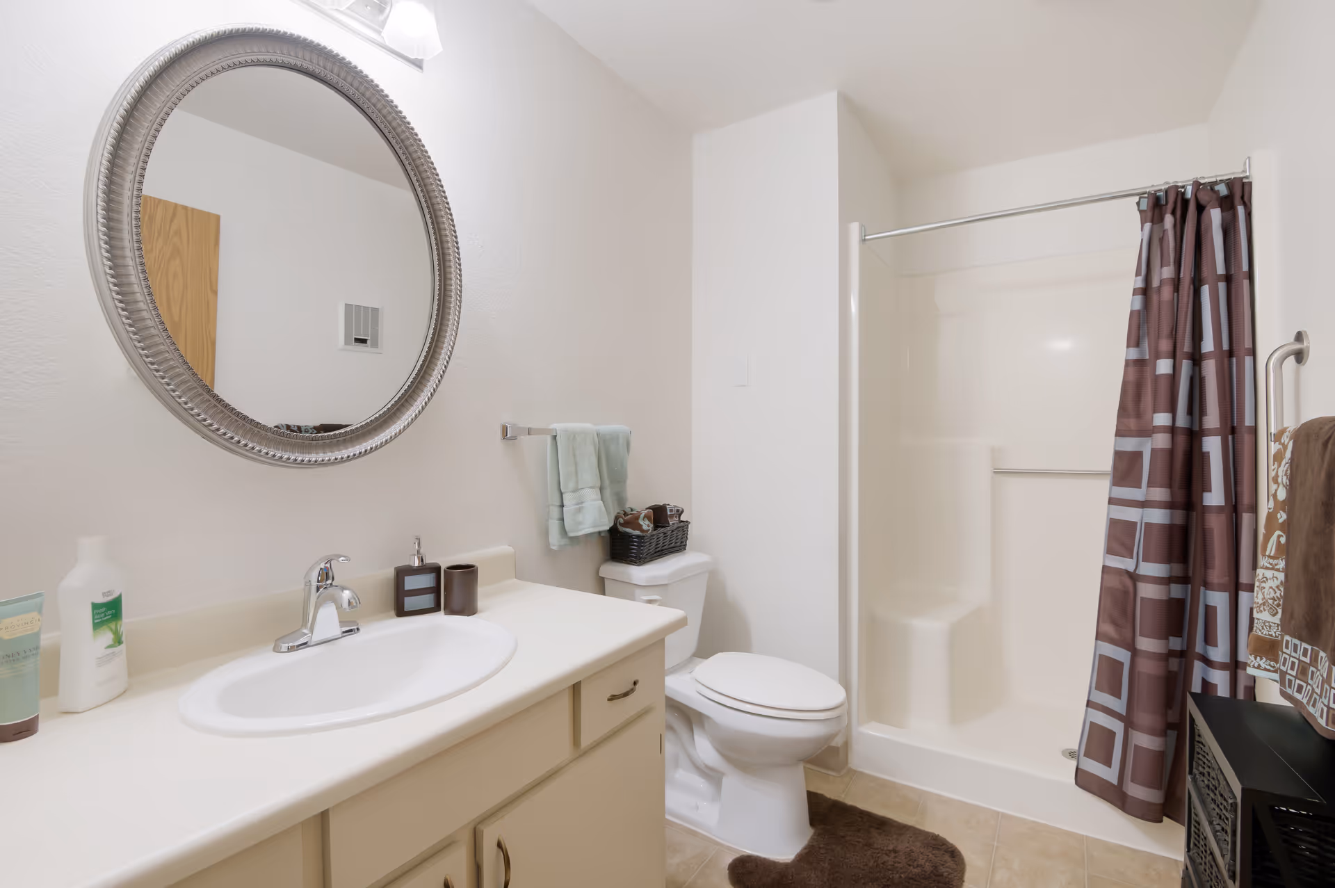 A clean bathroom with a white sink and countertop, a round silver-framed mirror above the sink, a toilet with a basket of towels on top, a shower with a brown and blue patterned curtain, and several towels hanging on racks.