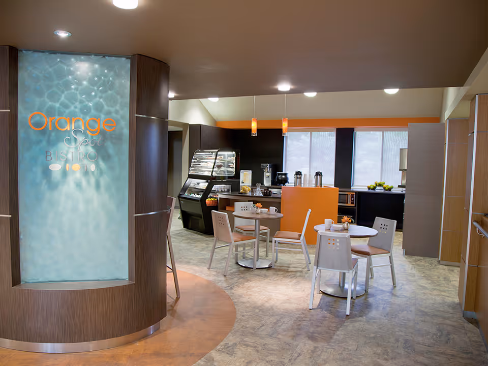 Interior bistro-style dining area with small round tables and chairs, a service counter, and a lit panel reading 'Orange Spot Bistro'.