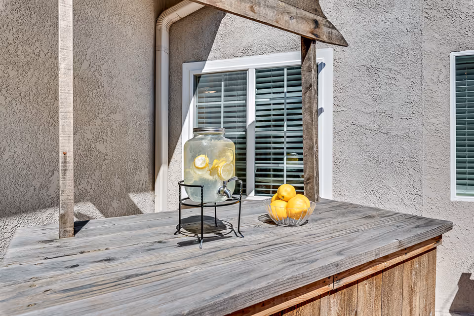 A wooden outdoor counter holding a glass lemon-water dispenser and a bowl of lemons in front of a stucco wall with shuttered windows.