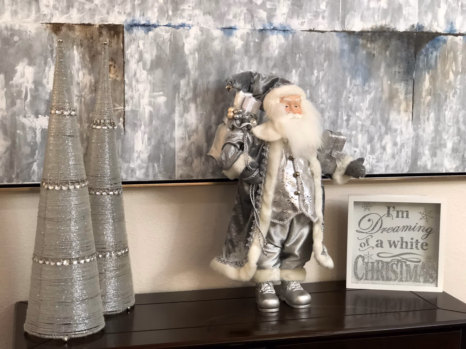 Silver-themed Christmas display featuring a Santa figurine, two cone trees, and a framed 'I'm Dreaming of a white Christmas' sign on a wooden console under abstract wall art.