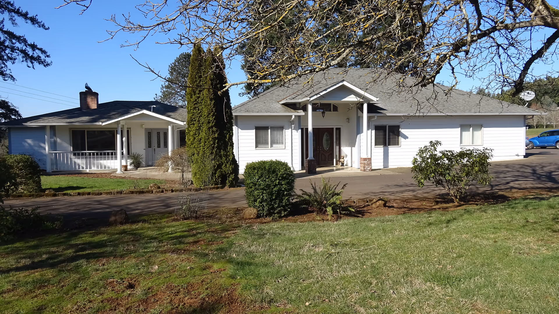 Exterior view of a single-story white building with a gray roof, surrounded by green grass and trees under a clear blue sky. The building has a covered entrance with two pillars and a driveway in front. There is a smaller adjacent building to the left with a chimney and a porch.