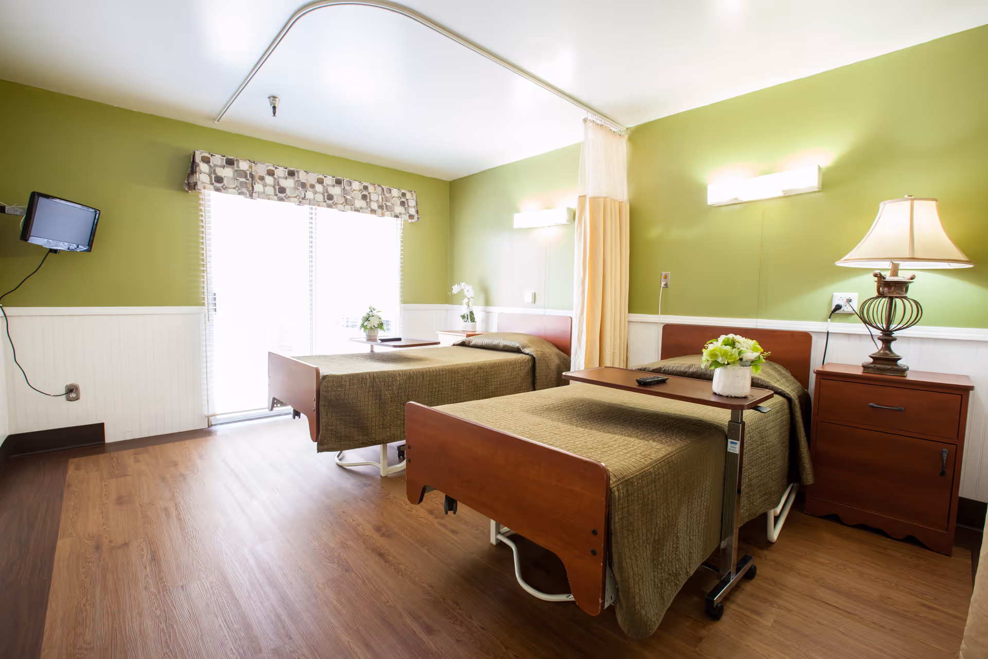 A bright nursing center bedroom with two hospital-style beds covered in brown bedspreads. Each bed has a small table with a flower vase on it. The room has green walls with white wainscoting, a wooden nightstand with a lamp, and a wall-mounted TV. Large windows with blinds and a patterned valance allow natural light to fill the room.