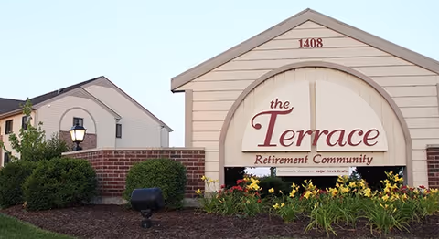 Exterior view of a retirement community sign reading 'the Terrace Retirement Community' with the address number 1408 above it, surrounded by landscaping with flowers and bushes, and a building and lamp post in the background.