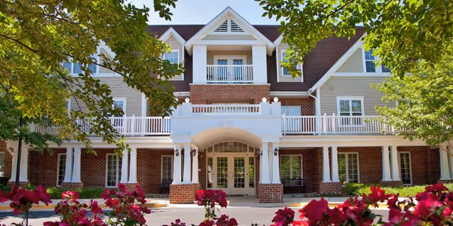 Front exterior view of a multi-story senior living facility named The Willows, featuring a brick and light-colored siding facade, white railings, a covered entrance, and surrounded by trees and flowering bushes.