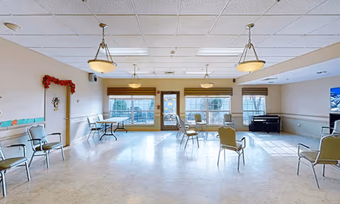 A spacious, well-lit common room with several chairs arranged around the perimeter and a few tables near the windows. The room has large windows with blinds, hanging ceiling lights, and a piano in the corner. The walls are light-colored, and there is a door decorated with a red garland on the left side.