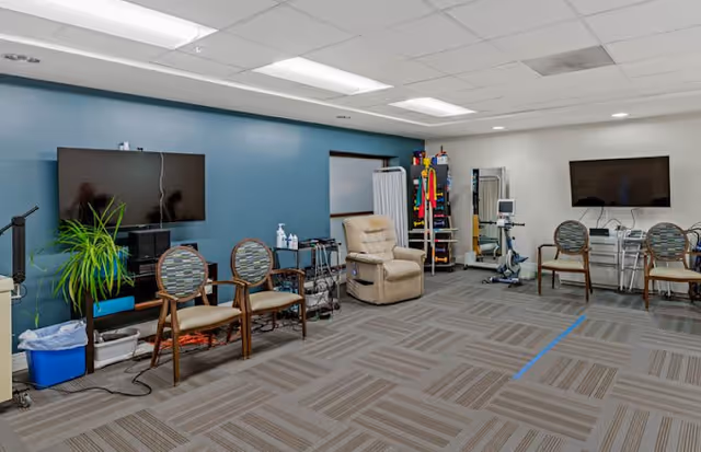 A spacious room with a blue accent wall featuring two mounted flat-screen TVs. The room contains several chairs with patterned cushions, a beige recliner, and various medical or therapy equipment including a walker, exercise machines, and a rack with colorful resistance bands. The floor is carpeted with a geometric pattern, and the ceiling has fluorescent lighting panels.