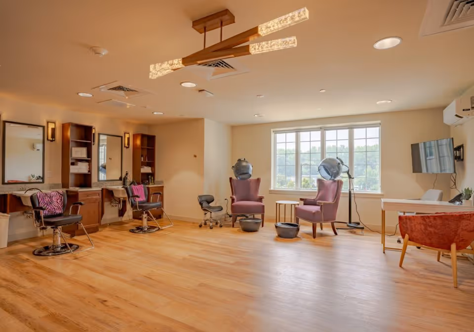 A spacious salon area with wooden flooring and beige walls. On the left side, there are two salon chairs in front of mirrors and shelves with towels and hair products. On the right side, two purple armchairs are positioned under hair dryers near a large window letting in natural light. A flat-screen TV is mounted on the wall, and a red chair is placed near a small desk.