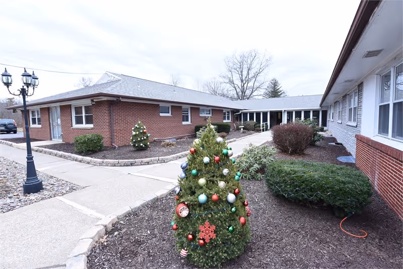 Outdoor courtyard area of a senior living facility with brick buildings on either side, a decorated small Christmas tree in the center, shrubs, and a lamppost on the left side. The sky is overcast.