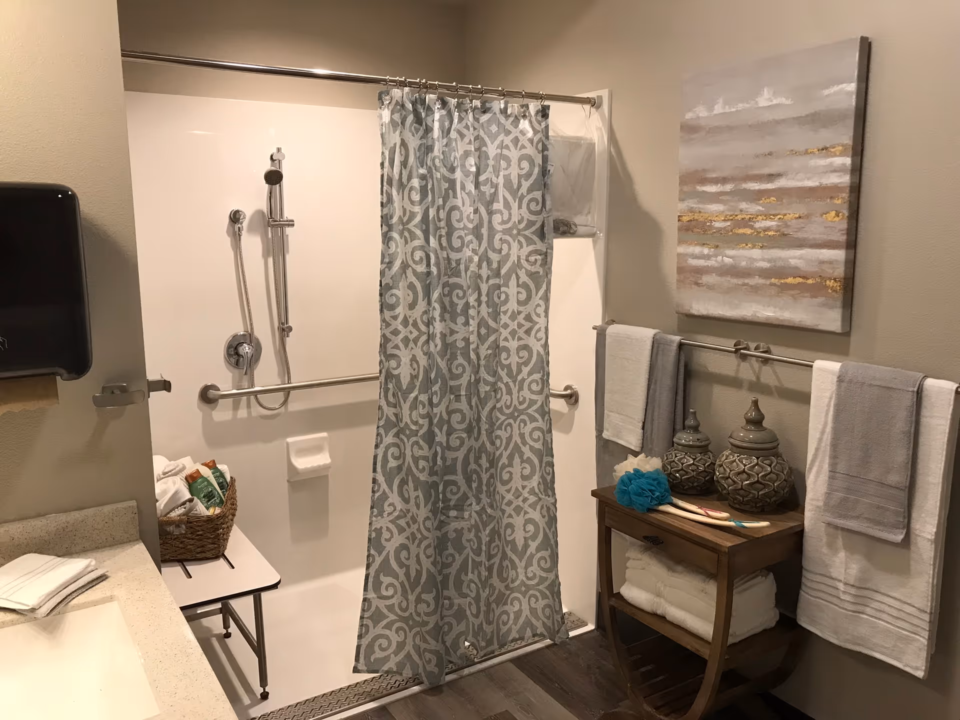 Accessible bathroom with a shower and patterned curtain, grab bars, towels, and a small wooden shelf holding toiletries and decorative jars.