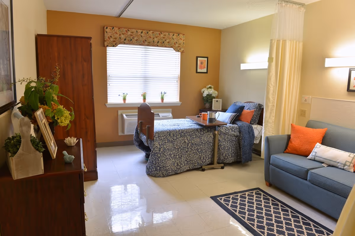 A cozy senior living bedroom with a single bed covered in a blue patterned bedspread, a small overbed table with a cup and pitcher, a blue couch with orange and white pillows, a wooden dresser with plants and picture frames, a window with blinds and a floral valance, and a privacy curtain partially drawn.