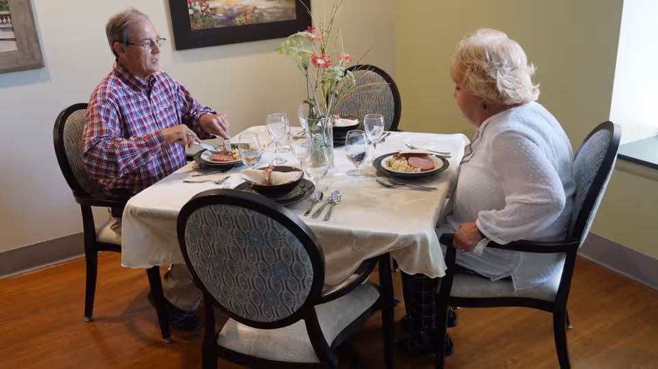Two elderly people sitting at a dining table in a well-lit room, eating a meal. The table is covered with a white tablecloth and set with plates, glasses, and silverware. A vase with flowers is placed in the center of the table. The room has wooden flooring and light-colored walls with framed artwork.