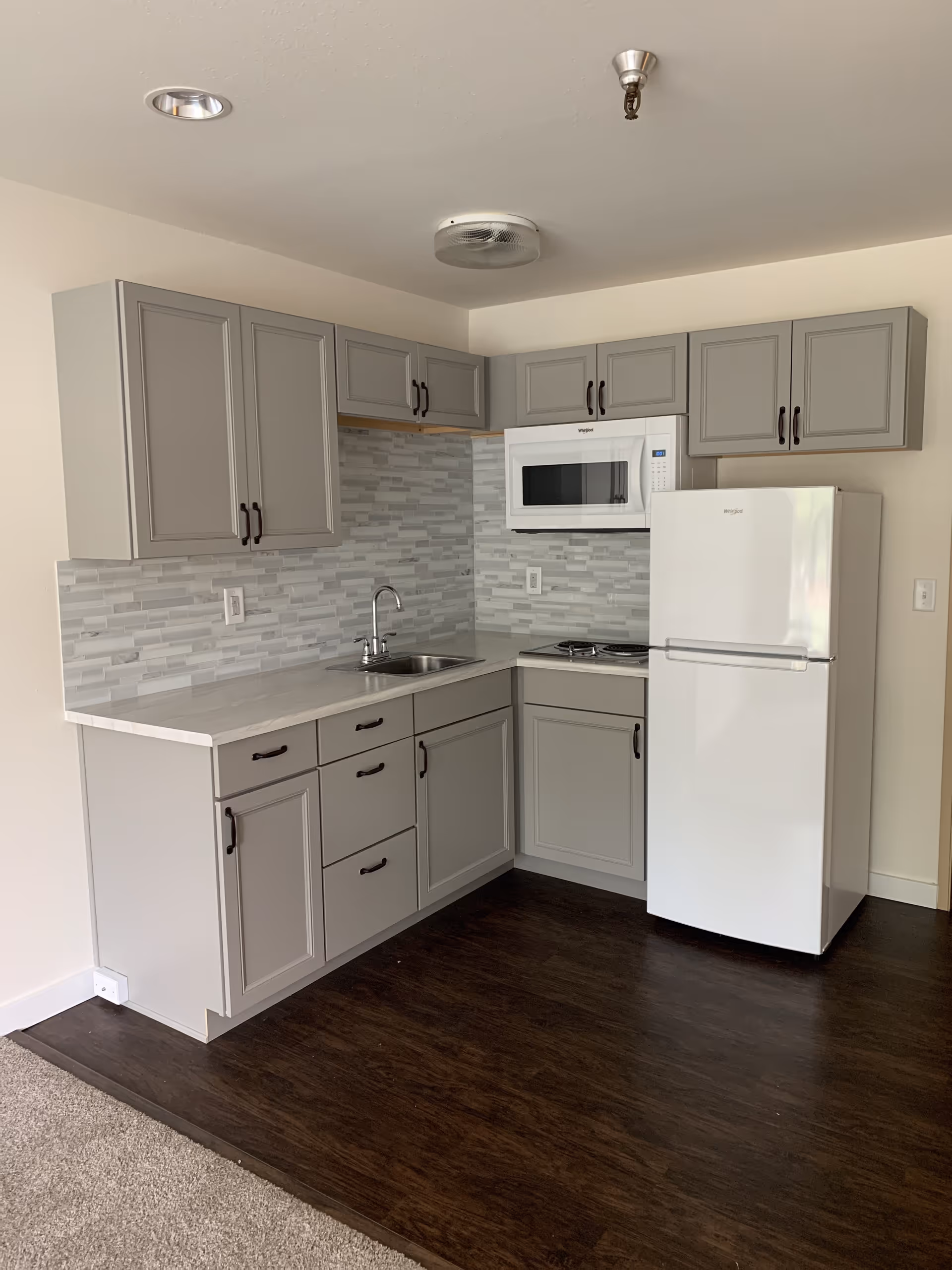 A small kitchen area with gray cabinets, a white countertop, a stainless steel sink, a white microwave mounted above a two-burner stove, and a white refrigerator. The backsplash features a gray and white tile pattern, and the floor is dark wood with carpet visible on the left side.