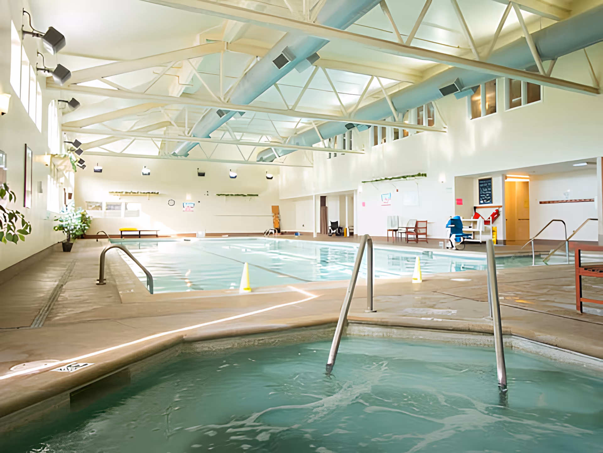 Indoor swimming pool area with a hot tub in the foreground and a larger pool in the background. The space has high ceilings with exposed beams and large windows letting in natural light. There are chairs and some plants along the walls.