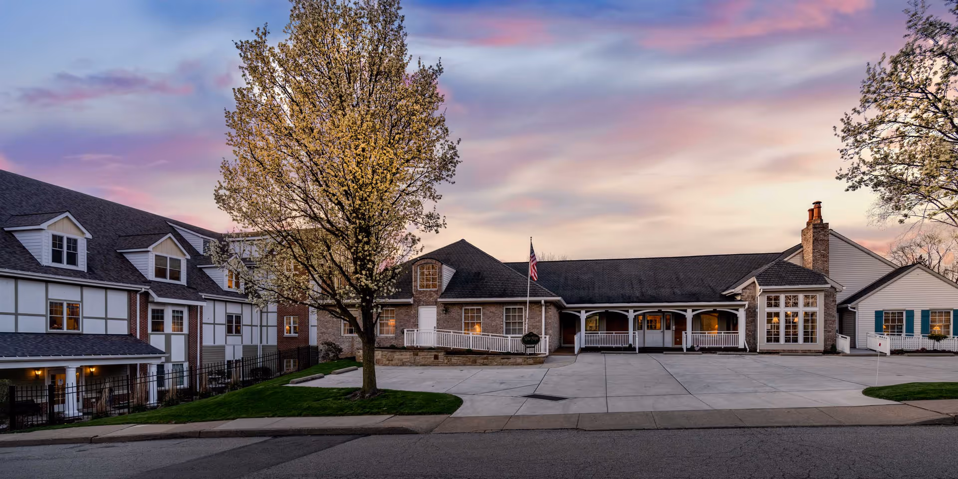Front view of a senior living facility building with a paved driveway, covered porch, and a flowering tree under a colorful sunset sky.