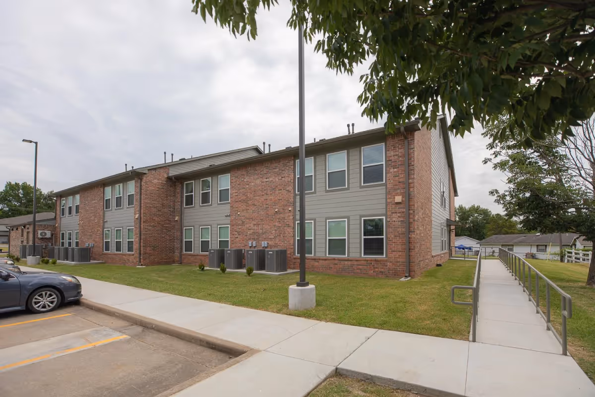 Exterior view of a two-story Hickory Crossing senior living building with brick and gray siding, a manicured lawn, and a ramp leading to the entrance.