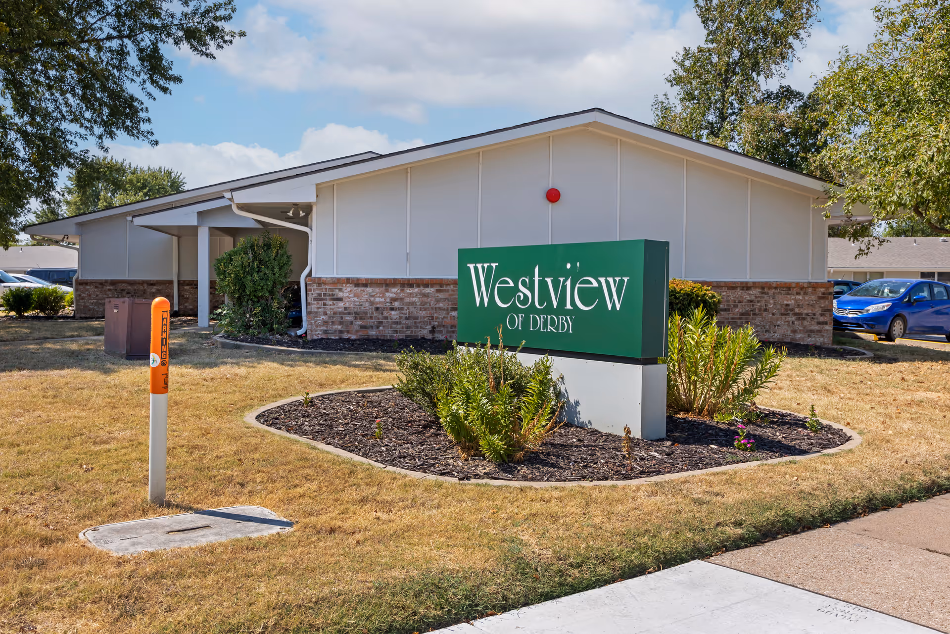Exterior front of the Westview of Derby building with a green sign in a mulched landscaped bed and a parking area nearby.