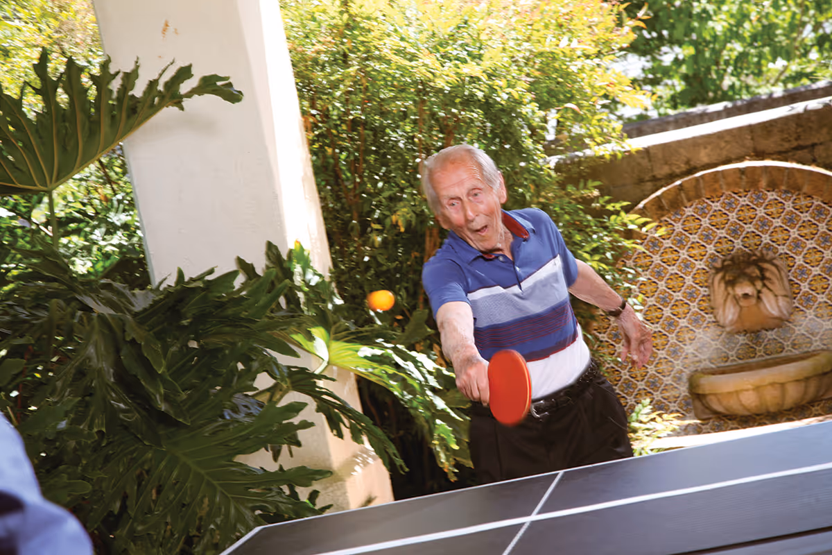 An elderly man playing table tennis outdoors near lush green plants and a decorative tiled wall with a lion head water fountain.