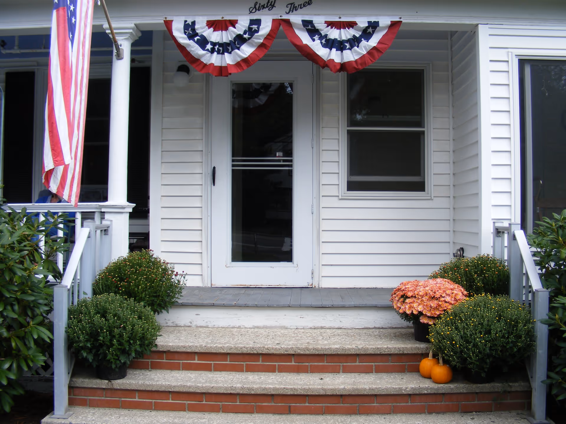 Front porch of a white house with steps leading up to a white door. The porch is decorated with American flag bunting, an American flag on a pole, potted green plants, orange flowers, and two small pumpkins.