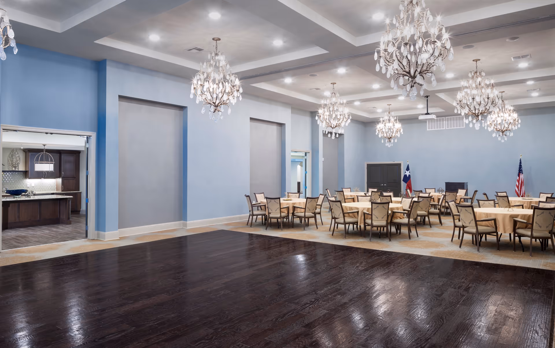 A spacious dining room with multiple round tables covered with beige tablecloths and surrounded by chairs. The room features light blue walls, dark wooden flooring in the foreground, and several ornate chandeliers hanging from a coffered ceiling. In the background, there are two flags, one of Texas and one of the United States, placed near a dark double door. To the left, an open doorway reveals a kitchen area with dark wooden cabinets and a granite countertop.