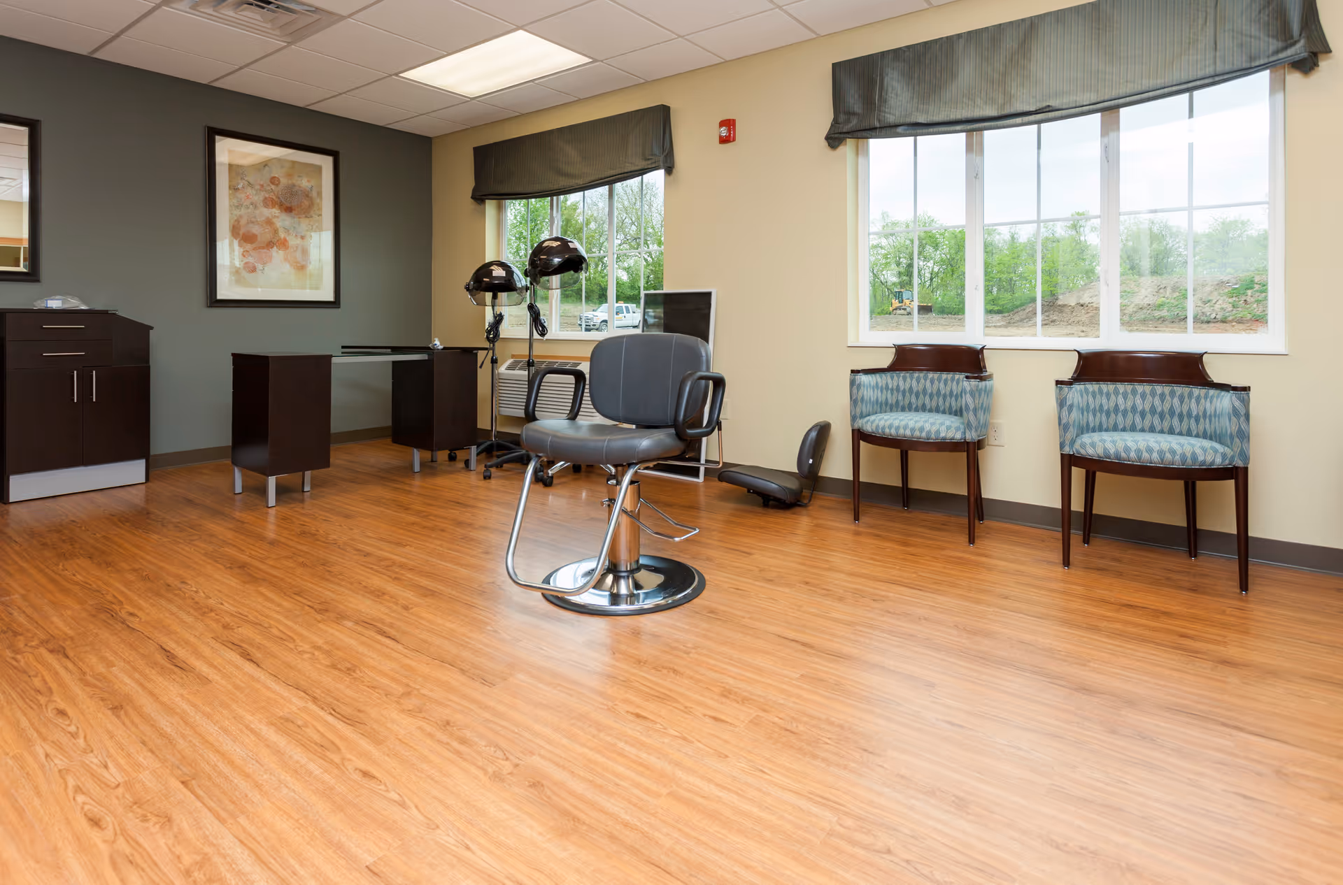 A bright room with wood flooring featuring a black salon chair in the center, two hair drying stations with black hooded dryers near a window, two patterned blue chairs against the wall, a dark wood cabinet, and a framed abstract artwork on the wall.
