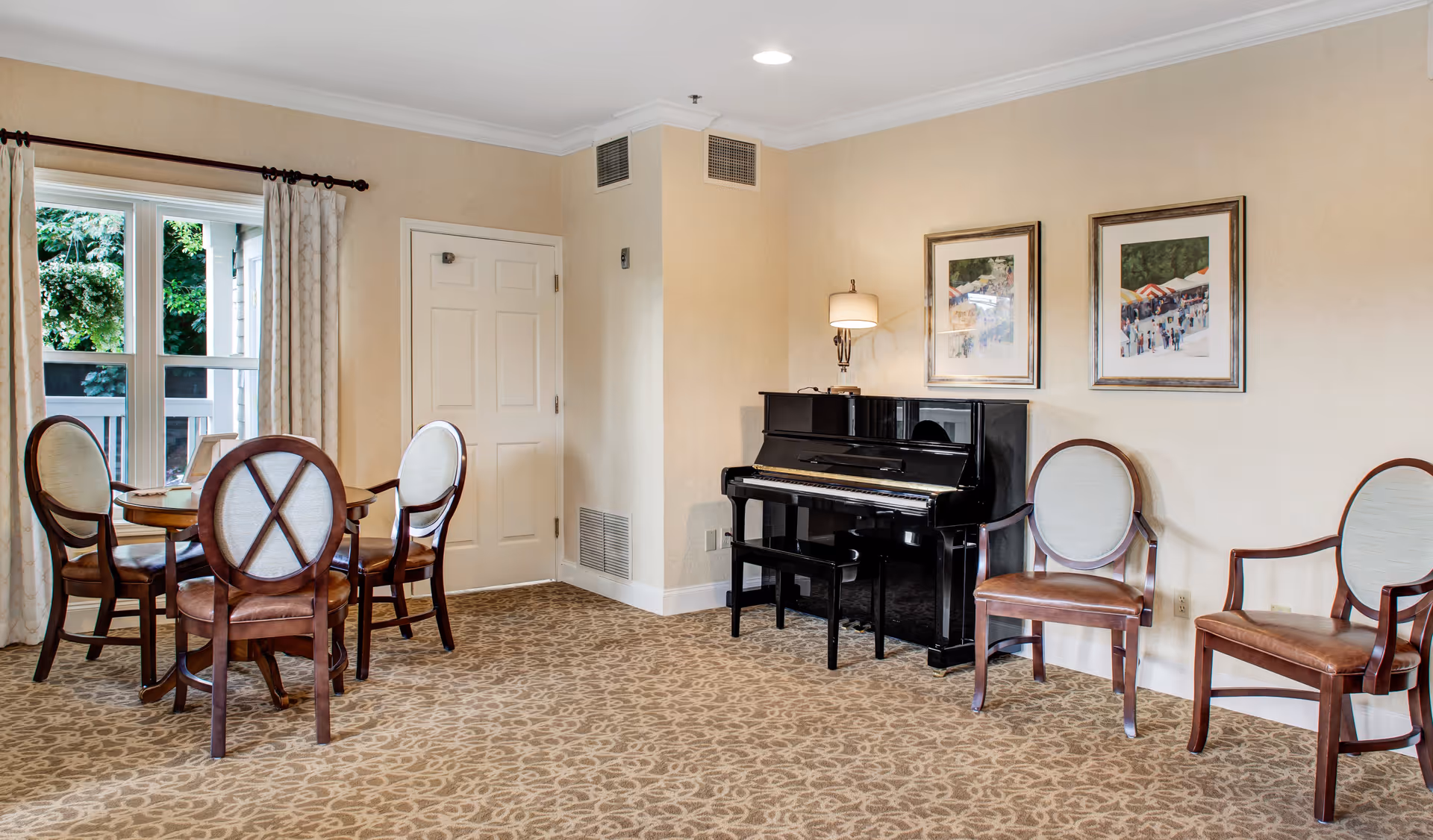 A cozy room with beige walls and patterned carpet featuring a black upright piano with a bench, two framed paintings above the piano, a table with four wooden chairs with cushioned seats near a window with curtains, and two additional wooden chairs with cushioned seats along the wall.