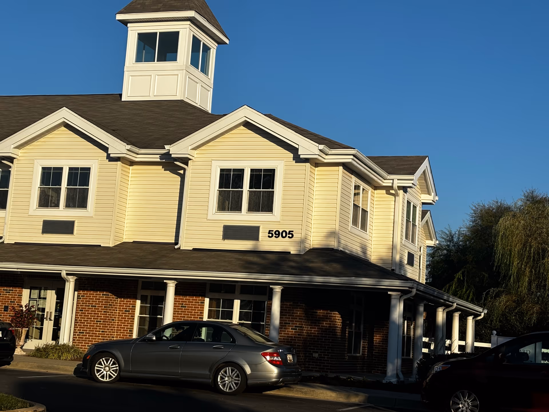 Front view of a two-story yellow building with a cupola, the address number "5905" on the facade, and cars parked in front.