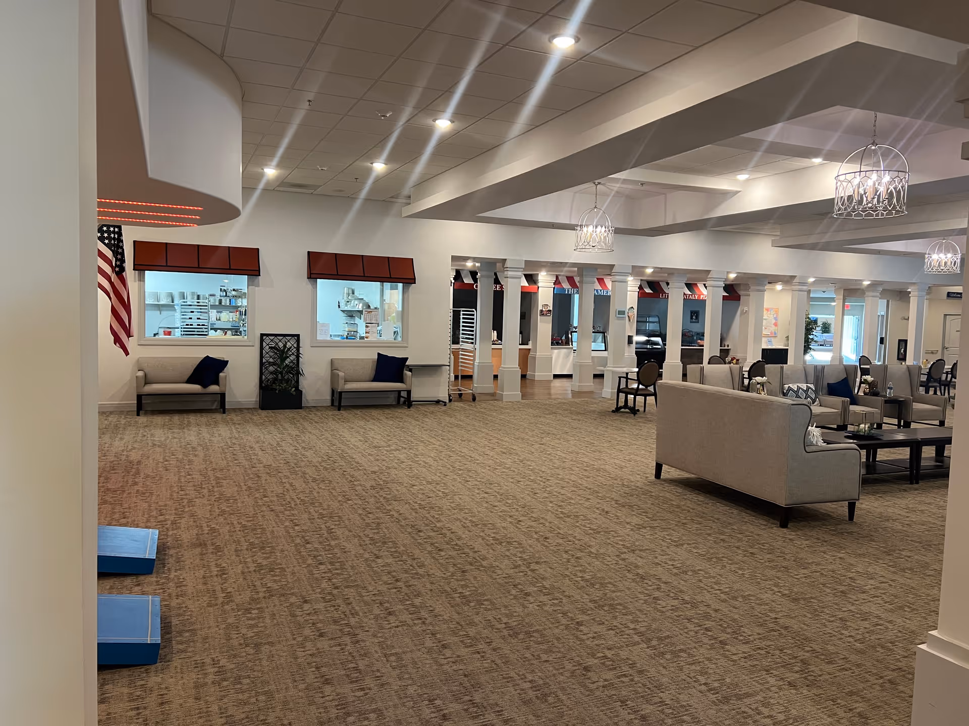 Spacious senior living facility common area with beige carpet, multiple seating arrangements including sofas and chairs, decorative hanging light fixtures, and an American flag on the left wall. There are windows showing a kitchen or food preparation area, and the room is well-lit with ceiling lights.