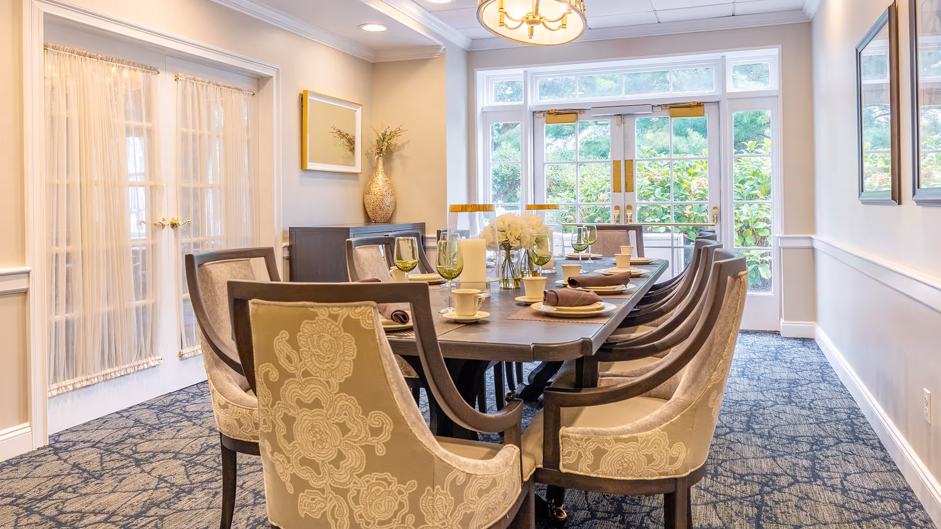 A well-lit dining room with a long wooden table set for a meal, featuring plates, cups, napkins, and green-tinted glasses. The room has upholstered chairs with floral patterns, a carpeted floor, large windows and glass doors letting in natural light, and decorative elements including a vase with flowers and framed artwork on the walls.