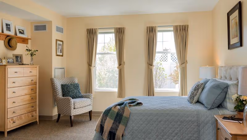Sunlit, neatly furnished bedroom with a made bed, upholstered armchair, wooden dresser, and two curtained windows.