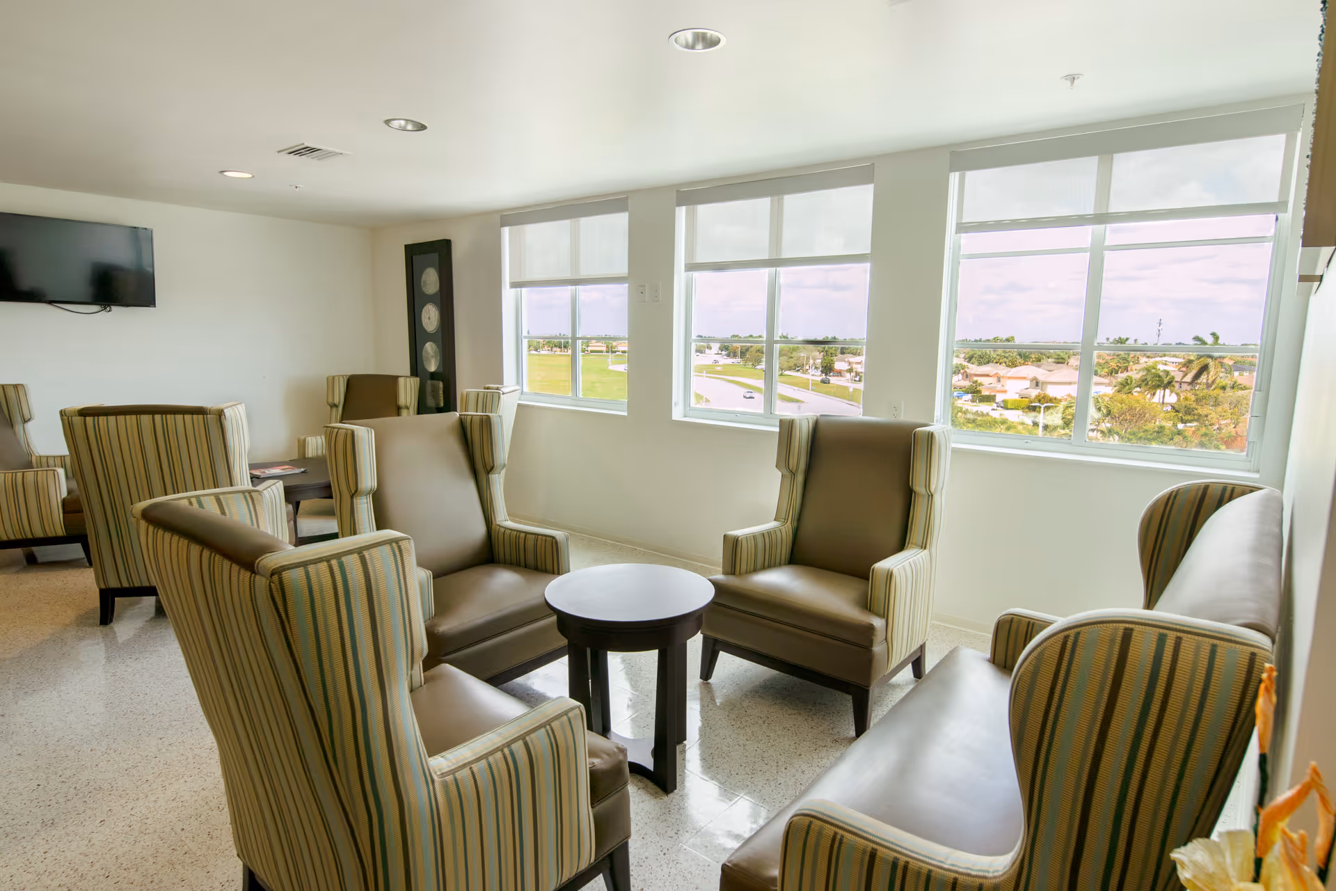 A bright lounge area with multiple striped armchairs and a small round table in the center. Large windows provide a view of a suburban neighborhood and greenery outside. A wall-mounted TV is visible on the left side of the room.