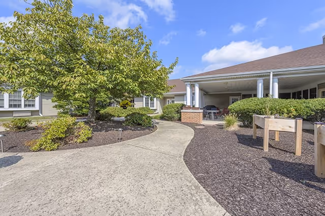 Curved concrete walkway through a landscaped courtyard leading to the covered entrance of a single-story senior living building under a blue sky.