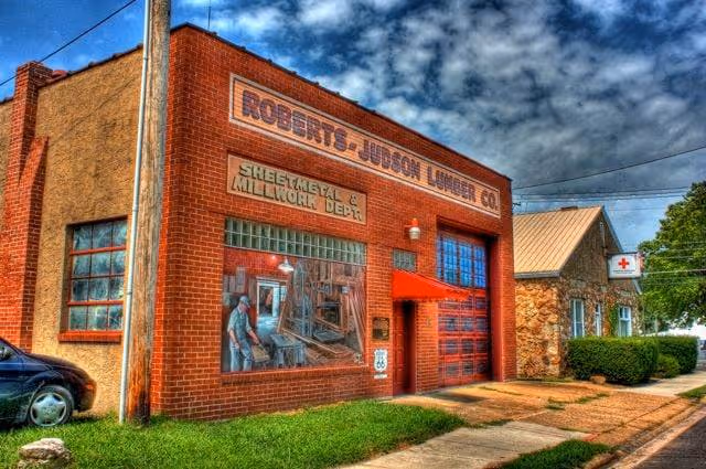 A red-brick storefront labeled 'Roberts-Judson Lumber Co.' with a painted mural, garage door and sidewalk under a dramatic cloudy sky.