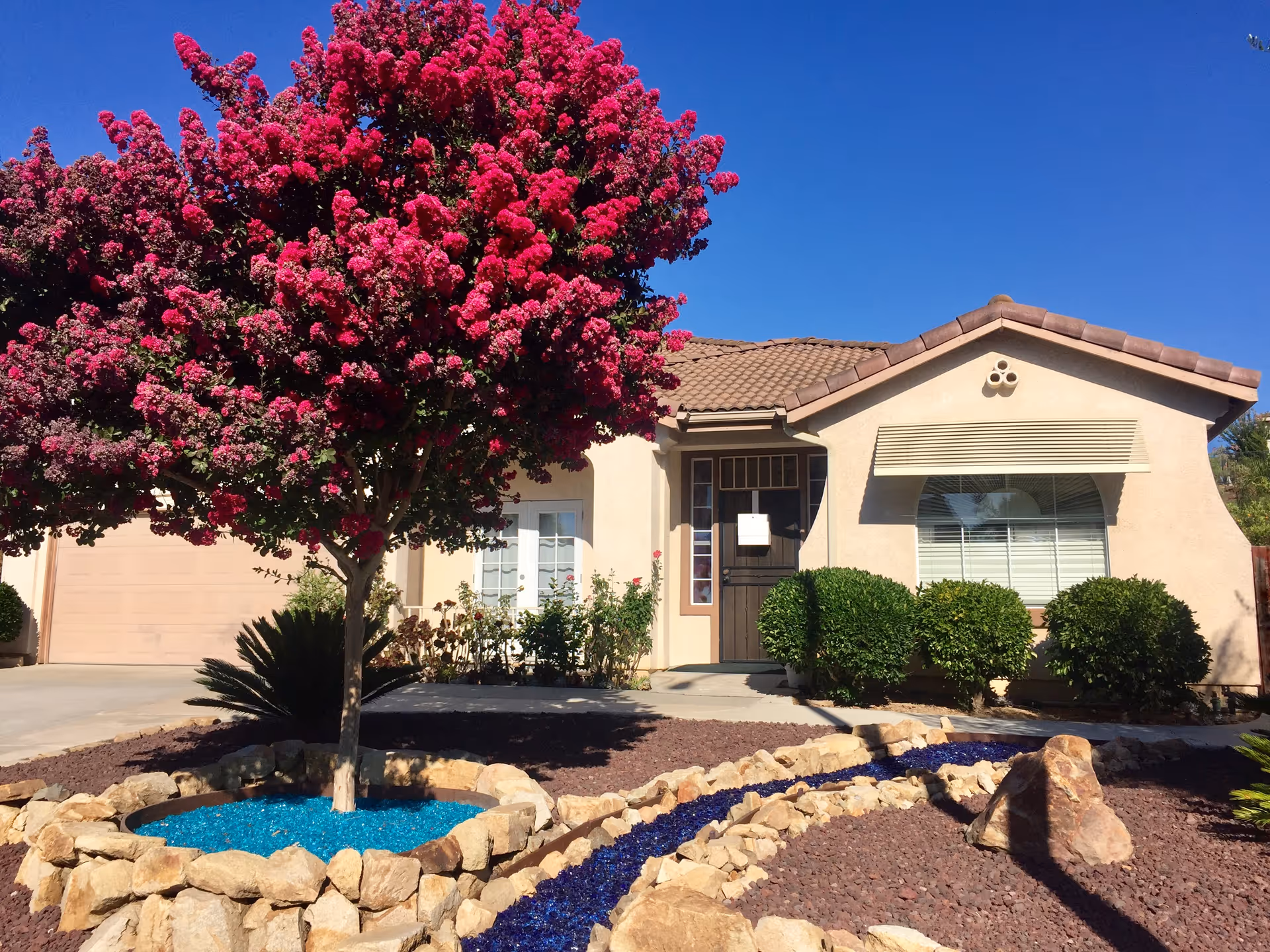 Front exterior view of a single-story house with a tiled roof, a large tree with bright pink flowers in the front yard, decorative blue and purple rocks bordered by stones, and neatly trimmed bushes near the windows and entrance.