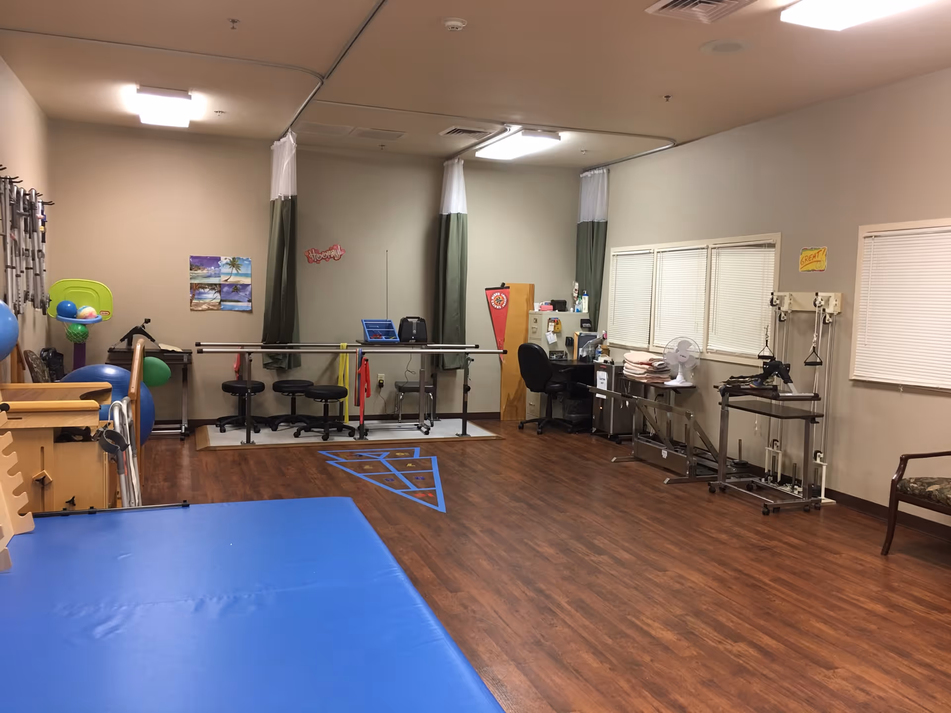 A rehabilitation or physical therapy room with wooden flooring and beige walls. The room contains various exercise and therapy equipment including parallel bars, stools, exercise balls, resistance bands, and pulley systems. There is a blue padded mat in the foreground, a desk with a chair and fan near the windows, and some colorful decorations on the walls.