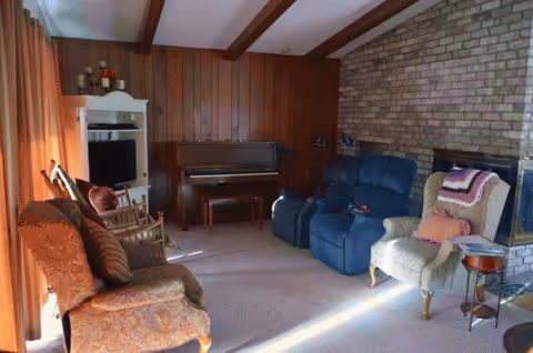 Cozy living room with wood paneling, brick fireplace, and vintage chairs and piano.