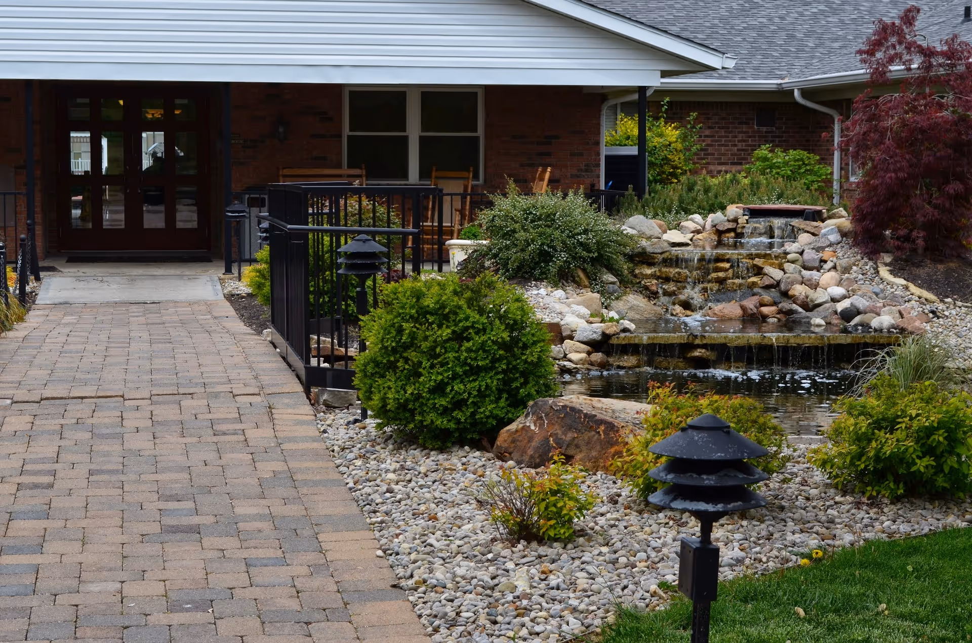 Paved walkway leading to a brick building entrance flanked by a landscaped rock garden and a small pond with a waterfall.