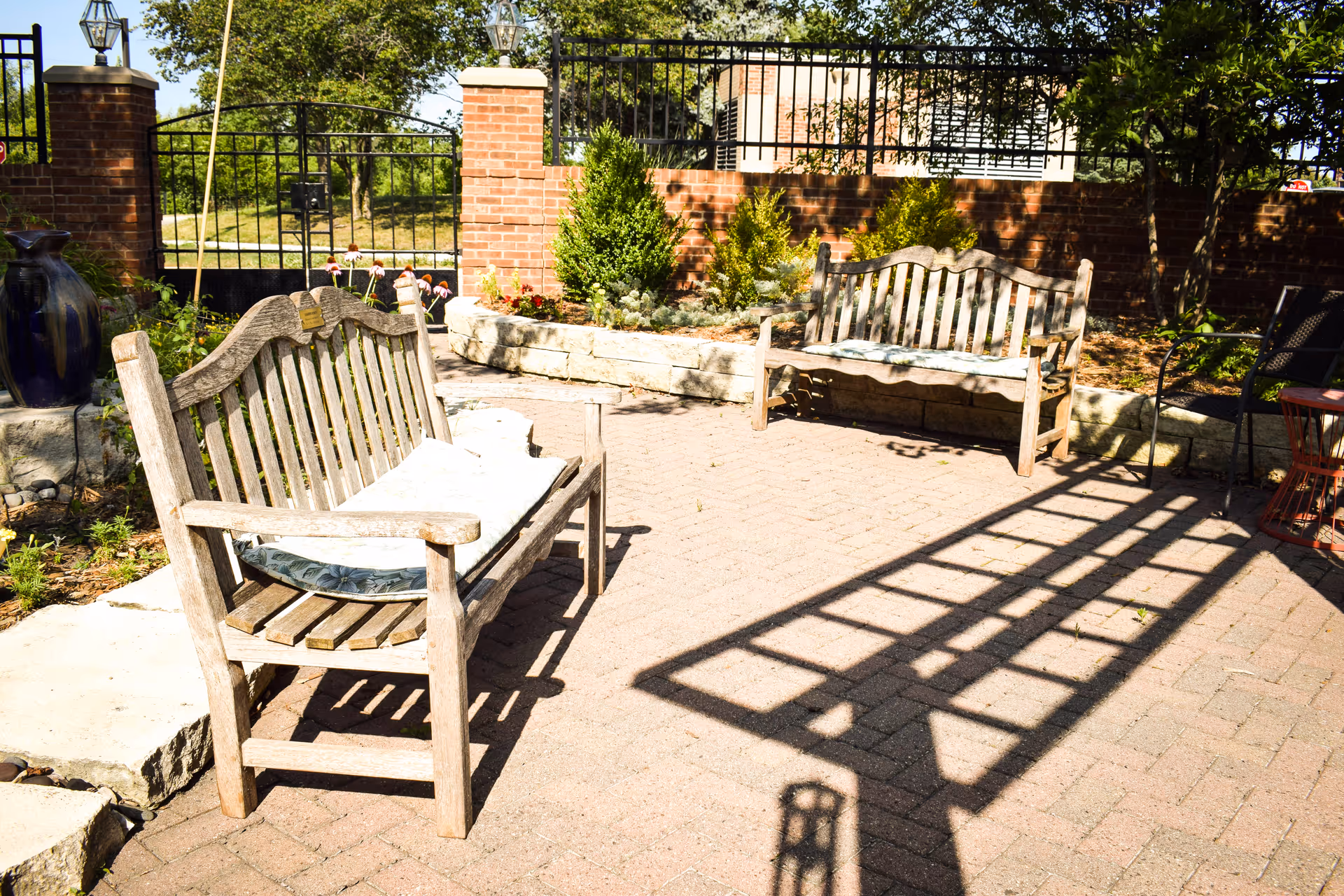 Outdoor patio area with two wooden benches, some cushions on the benches, brick pillars with lanterns, a black metal gate, and a garden with plants and trees in the background.