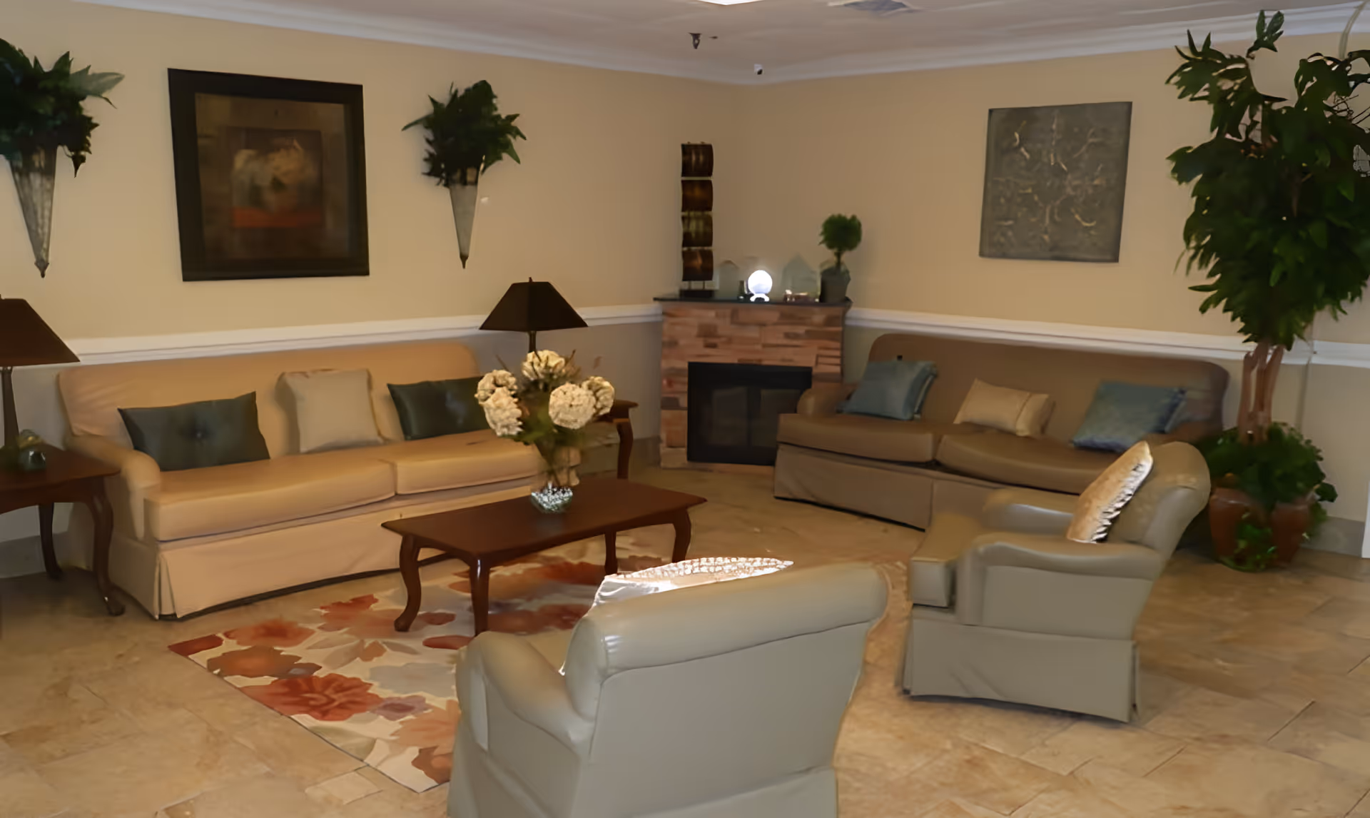 A cozy living room with two beige sofas, two beige armchairs, a wooden coffee table with a floral arrangement, a fireplace with decorative items on the mantel, wall art, and several green plants. The room has beige walls and tiled flooring with a floral area rug.